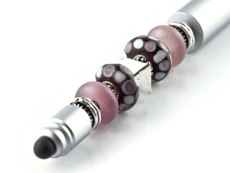 May include: A silver stylus pen with a black tip and a series of pink and purple glass beads with white polka dots.