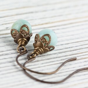 May include: A pair of earrings featuring pale blue, spherical beads. The beads are accented with ornate, bronze-colored metalwork. The earrings have a simple, curved hook design, and are displayed on a white, wooden surface.