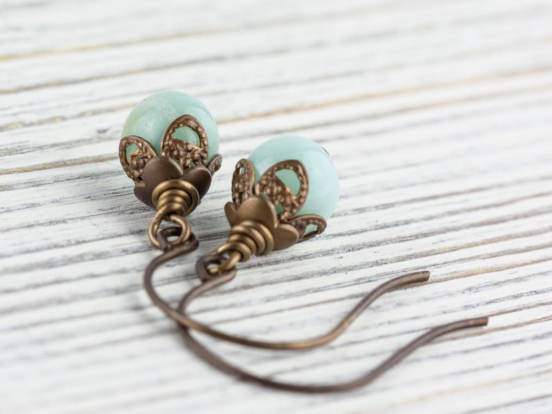 May include: A pair of earrings featuring pale blue, spherical beads. The beads are accented with ornate, bronze-colored metalwork. The earrings have a simple, curved hook design, and are displayed on a white, wooden surface.