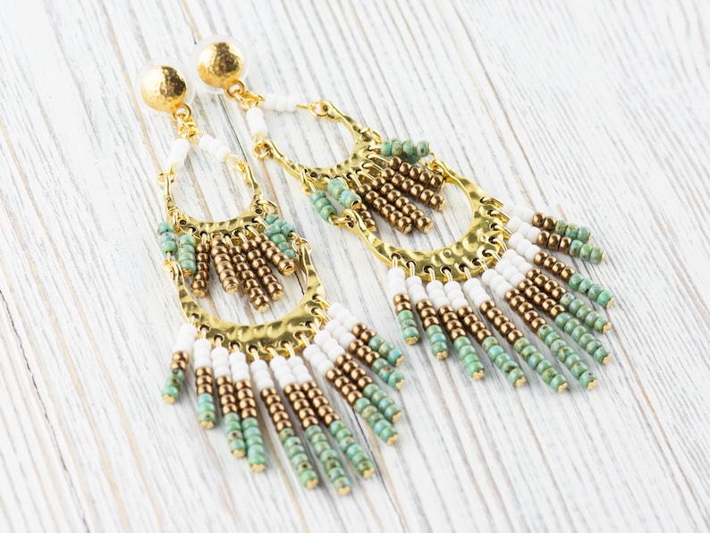 May include: A pair of long, gold-toned earrings with white, green, and brown beads. The earrings have a hammered metal design and dangle from the earlobe.