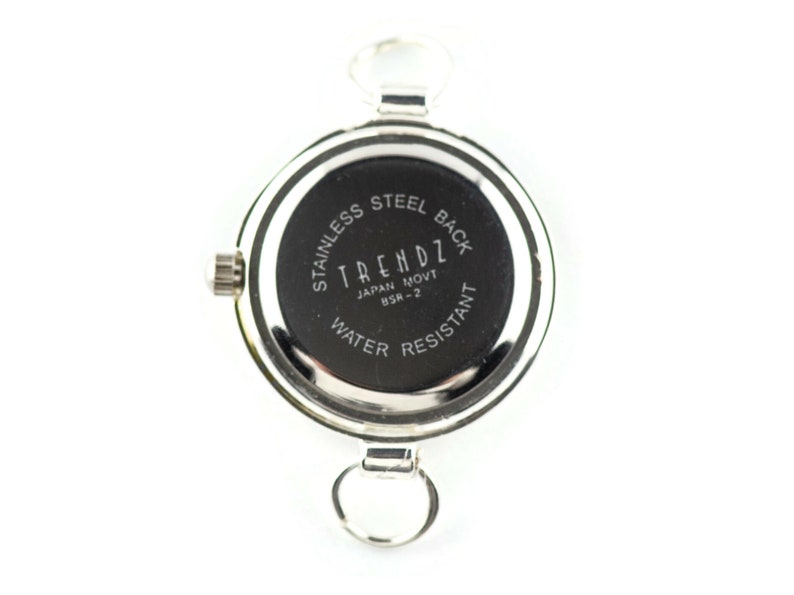 May include: A silver watch with a black face. The watch back reads "TRENDZ JAPAN MOVT BSR-2 WATER RESISTANT STAINLESS STEEL BACK." It has a circular design, a crown, and two loops for attachment.