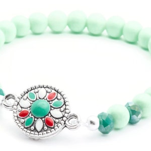 May include: A mint green beaded bracelet with a silver charm featuring a green, red and white floral design.