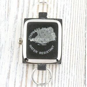 May include: A square Geneva watch with a stainless steel back, water resistant, and Japan movement. The watch is attached to a keychain with two silver rings. The watch face is black with silver trim.
