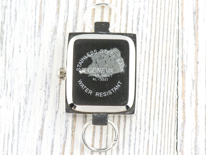 May include: A square Geneva watch with a stainless steel back, water resistant, and Japan movement. The watch is attached to a keychain with two silver rings. The watch face is black with silver trim.