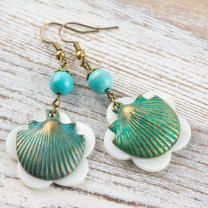 May include: A pair of earrings featuring a turquoise bead and a shell-shaped charm. The shell is made of metal and has a green and gold finish. The earrings are hanging against a white wooden background.