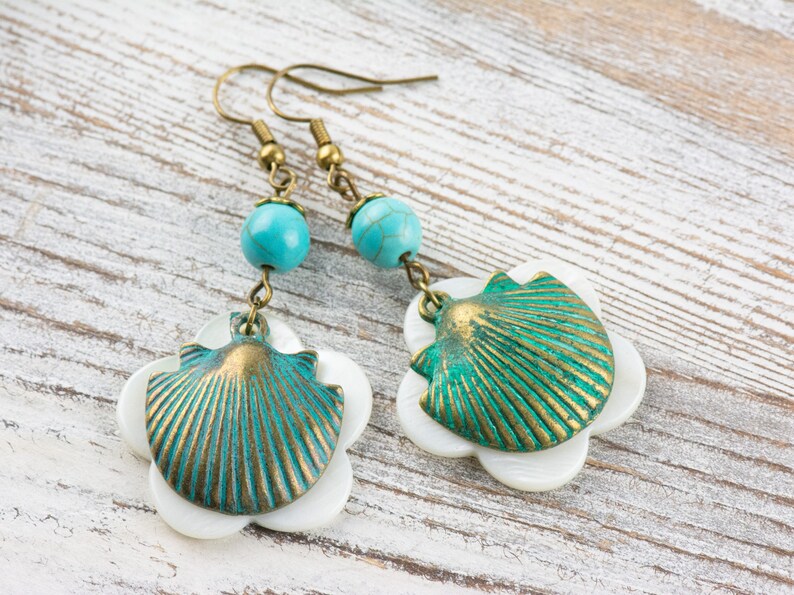 May include: A pair of earrings featuring a turquoise bead and a shell-shaped charm. The shell is made of metal and has a green and gold finish. The earrings are hanging against a white wooden background.