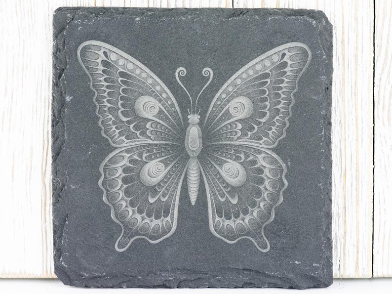 Puede incluir: Un posavasos de pizarra negra con un dise&ntilde;o de mariposa grabado en blanco. La mariposa tiene detalles intrincados y una apariencia delicada.