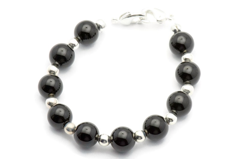 Puede incluir: Una pulsera con grandes cuentas negras brillantes intercaladas con cuentas m&aacute;s peque&ntilde;as de color plateado. La pulsera se asegura con un cierre plateado. Las cuentas est&aacute;n espaciadas uniformemente, creando un dise&ntilde;o simple pero elegante.