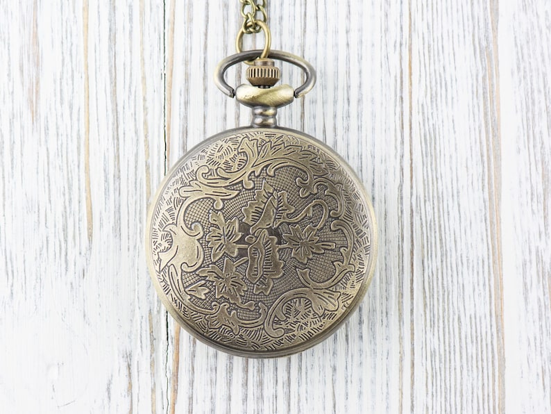 Pode incluir: Um rel&oacute;gio de bolso dourado com um design floral decorativo no mostrador. O rel&oacute;gio est&aacute; pendurado em uma corrente.