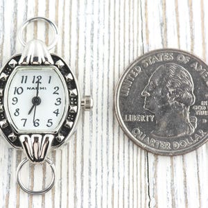 May include: A silver pocket watch with a white face and black numerals, next to a quarter. The watch has a decorative bezel and a ring for attachment. The quarter is a United States quarter dollar.