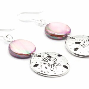 Puede incluir: Un par de pendientes de plata con una concha iridiscente rosa y un dije de d&oacute;lar de arena plateado.
