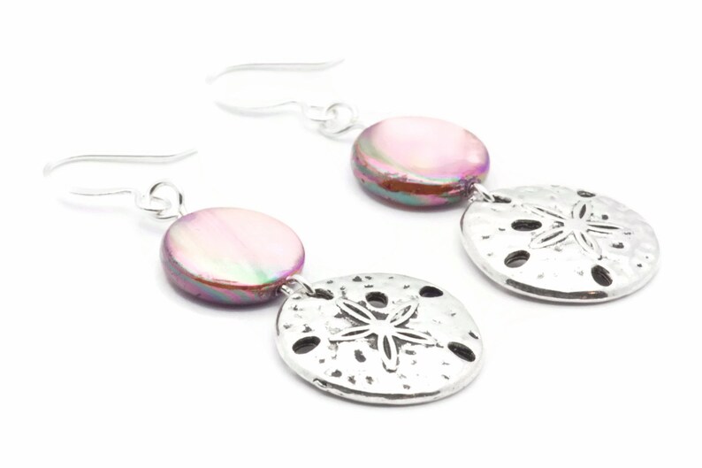 Puede incluir: Un par de pendientes de plata con una concha iridiscente rosa y un dije de d&oacute;lar de arena plateado.