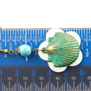 May include: A pair of earrings featuring a turquoise bead and a shell charm. The shell charm is made of metal and has a green and gold finish. The earrings are measured against a ruler.