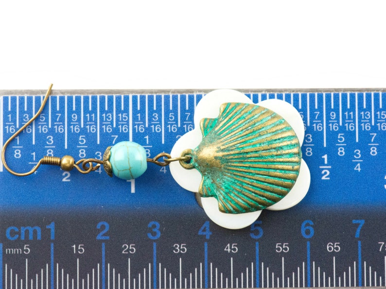 May include: A pair of earrings featuring a turquoise bead and a shell charm. The shell charm is made of metal and has a green and gold finish. The earrings are measured against a ruler.