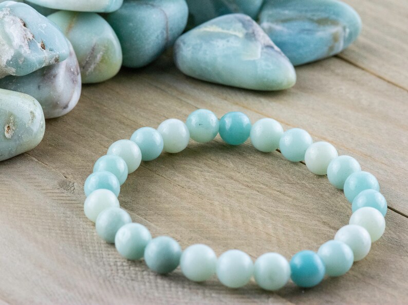 May include: A light blue gemstone bracelet made of round beads. The bracelet is on a light brown wooden surface. There are several loose light blue gemstones in the background.
