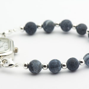 May include: A silver watch with a square face and a blue agate bead bracelet. The watch face has black Roman numeral hour markers and black hands.