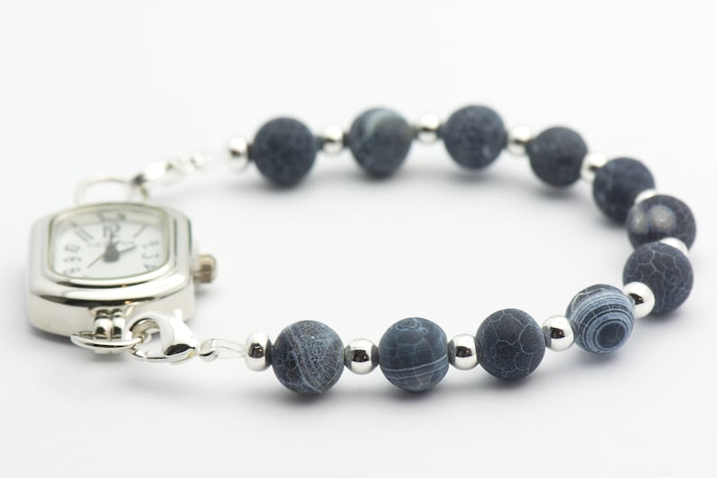May include: A silver watch with a square face and a blue agate bead bracelet. The watch face has black Roman numeral hour markers and black hands.