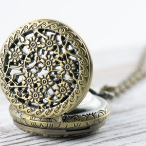 K&ouml;nnte beinhalten: Eine antike Taschenuhr mit einem durchbrochenen Design. Das Uhrengeh&auml;use hat eine bronzefarbene Oberfl&auml;che, und das offene Zifferblatt zeigt das Innenleben. Eine Kette ist an der Uhr befestigt.