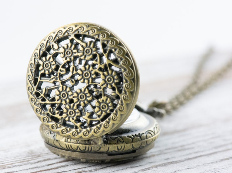 K&ouml;nnte beinhalten: Eine antike Taschenuhr mit einem durchbrochenen Design. Das Uhrengeh&auml;use hat eine bronzefarbene Oberfl&auml;che, und das offene Zifferblatt zeigt das Innenleben. Eine Kette ist an der Uhr befestigt.