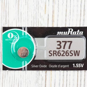 May include: A silver oxide battery with the text "muRata 377 SR626SW" and "Silver Oxide Oxyde d'argent 1.55V" printed on the packaging. The battery is 0% mercury.