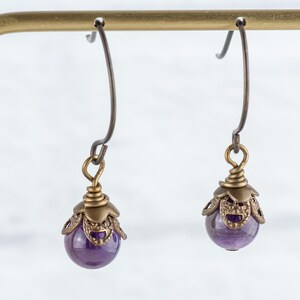 K&ouml;nnte beinhalten: Ein Paar Ohrh&auml;nger mit einer tiefvioletten, kugelf&ouml;rmigen Perle. Die Perle ist in einer verzierten, antik-messingfarbenen Metallkappe eingeschlossen und an einem gebogenen Ohrhaken aufgeh&auml;ngt. Die Ohrringe werden vor einem neutralen Hintergrund pr&auml;sentiert.