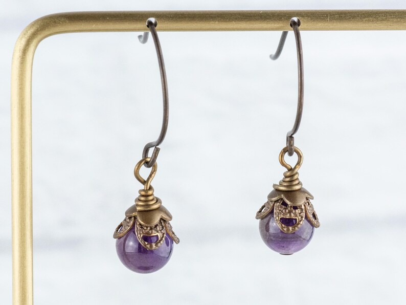 K&ouml;nnte beinhalten: Ein Paar Ohrh&auml;nger mit einer tiefvioletten, kugelf&ouml;rmigen Perle. Die Perle ist in einer verzierten, antik-messingfarbenen Metallkappe eingeschlossen und an einem gebogenen Ohrhaken aufgeh&auml;ngt. Die Ohrringe werden vor einem neutralen Hintergrund pr&auml;sentiert.