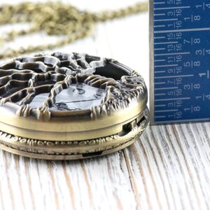 May include: An antique bronze pocket watch with a tree design on the cover. The watch is approximately 4.4 cm in diameter. A ruler is next to the watch, showing the measurement.