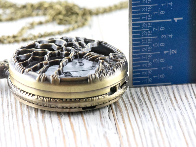May include: An antique bronze pocket watch with a tree design on the cover. The watch is approximately 4.4 cm in diameter. A ruler is next to the watch, showing the measurement.