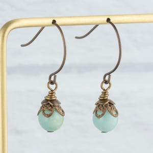 May include: A pair of dangle earrings with a light teal, round bead. The beads are topped with a bronze-colored filigree cap and hang from bronze-colored ear wires. The earrings are displayed on a gold-colored earring stand.
