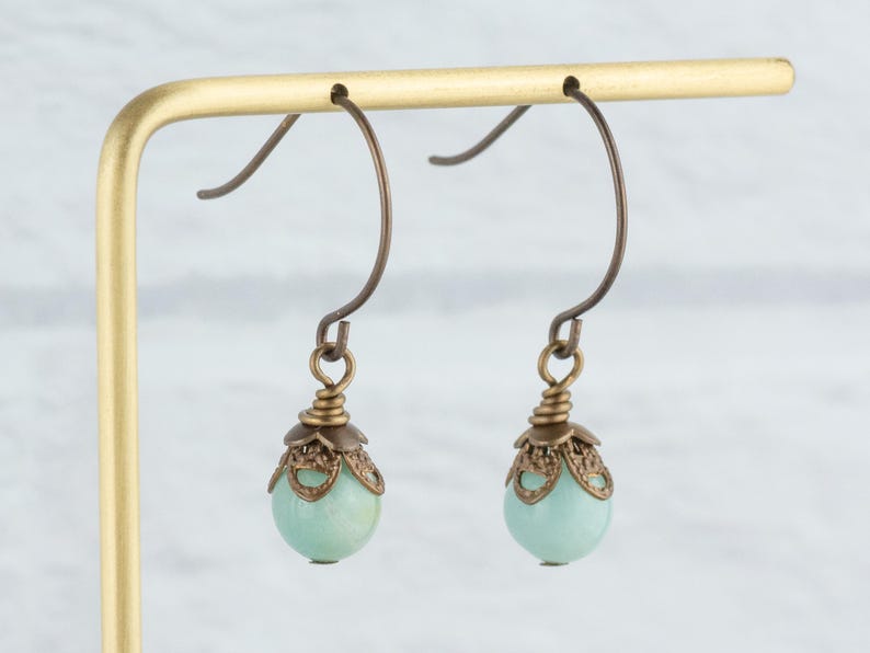 May include: A pair of dangle earrings with a light teal, round bead. The beads are topped with a bronze-colored filigree cap and hang from bronze-colored ear wires. The earrings are displayed on a gold-colored earring stand.