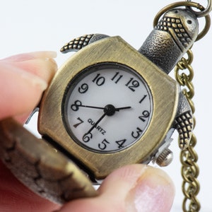 May include: A gold-toned pocket watch shaped like a turtle with a white face and black hands. The watch has a chain attached to it and says "Quartz" on the face.