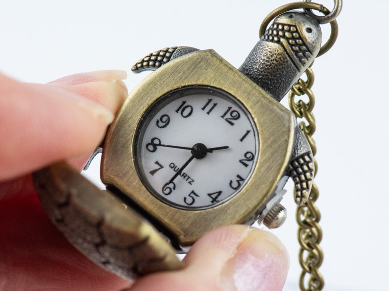 May include: A gold-toned pocket watch shaped like a turtle with a white face and black hands. The watch has a chain attached to it and says "Quartz" on the face.