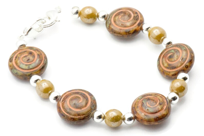 May include: A bracelet with large, round, brown and green glass beads, each with a spiral pattern. It also features smaller olive-green beads and silver-coloured spacer beads. A silver-coloured clasp secures the bracelet.