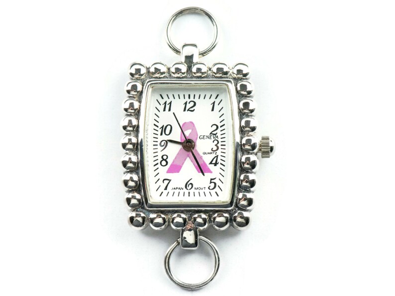 May include: Silver-toned rectangular pocket watch with a white face featuring black numerals and a pink ribbon. The watch has a beaded border and two ring attachments. The watch face reads "GENEVA QUARTZ JAPAN MOVT".
