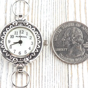 Op de afbeelding: Een zilverkleurig zakhorloge met een witte wijzerplaat en zwarte cijfers, naast een Amerikaanse kwart dollar. Het horloge heeft een decoratieve rand en de merknaam "NARMI" is zichtbaar. Het horloge heeft een diameter van ongeveer 3,8 cm.