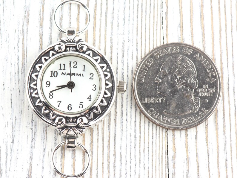 Op de afbeelding: Een zilverkleurig zakhorloge met een witte wijzerplaat en zwarte cijfers, naast een Amerikaanse kwart dollar. Het horloge heeft een decoratieve rand en de merknaam "NARMI" is zichtbaar. Het horloge heeft een diameter van ongeveer 3,8 cm.