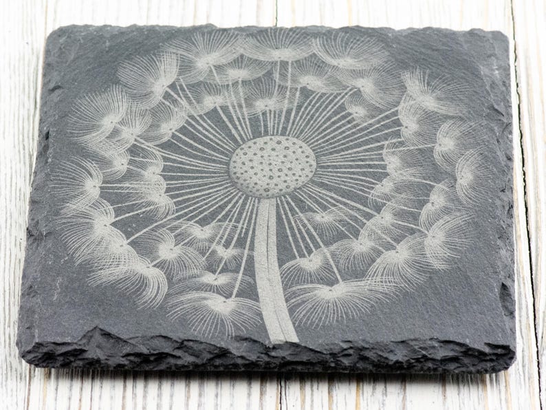 K&ouml;nnte beinhalten: Ein schwarzer Schieferuntersetzer mit einem wei&szlig;en, ge&auml;tzten Design eines L&ouml;wenzahns.