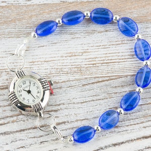 May include: A silver watch with a white face and black numbers is attached to a blue beaded bracelet. The bracelet is made of oval-shaped beads with silver spacers.