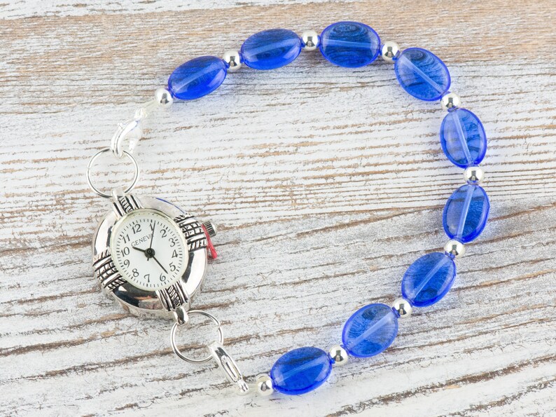 May include: A silver watch with a white face and black numbers is attached to a blue beaded bracelet. The bracelet is made of oval-shaped beads with silver spacers.