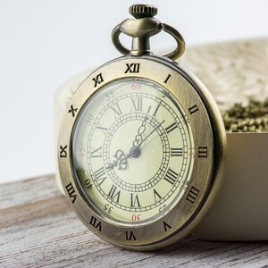 Op de afbeelding: Antieke messing zakhorloge met een cr&egrave;mekleurige wijzerplaat en zwarte Romeinse cijfers. Het horloge heeft een doorzichtige glazen afdekking en een kleine lus aan de bovenkant voor een ketting. Het horloge staat tegen een neutrale achtergrond.