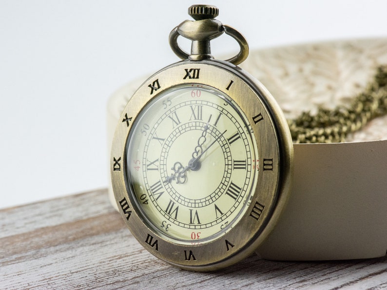 Op de afbeelding: Antieke messing zakhorloge met een cr&egrave;mekleurige wijzerplaat en zwarte Romeinse cijfers. Het horloge heeft een doorzichtige glazen afdekking en een kleine lus aan de bovenkant voor een ketting. Het horloge staat tegen een neutrale achtergrond.
