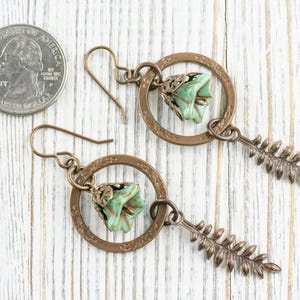 May include: A pair of handcrafted earrings featuring a bronze-coloured circular frame with a green flower and a fern-shaped charm. The earrings are displayed next to a coin for size reference.