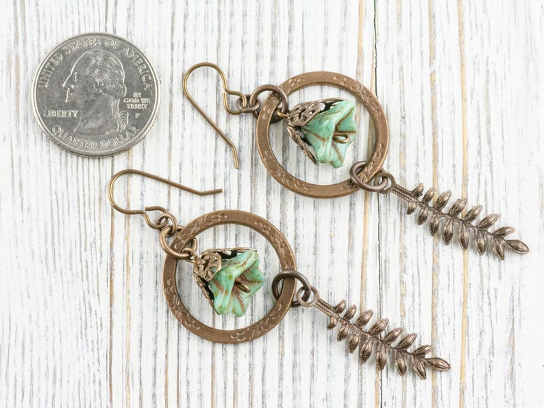 May include: A pair of handcrafted earrings featuring a bronze-coloured circular frame with a green flower and a fern-shaped charm. The earrings are displayed next to a coin for size reference.