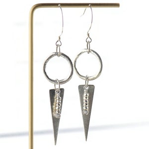 May include: Silver dangle earrings with a circle and triangle design. The triangle features a row of small silver beads and a clear stone.