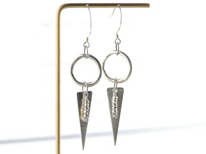 May include: Silver dangle earrings with a circle and triangle design. The triangle features a row of small silver beads and a clear stone.
