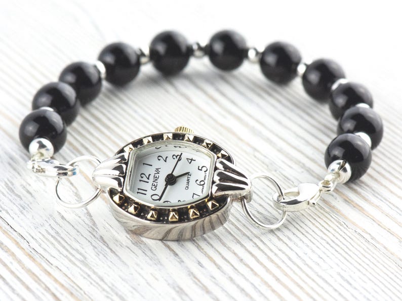 Peut inclure: Une montre Geneva &agrave; quartz avec un bo&icirc;tier argent&eacute; et un cadran blanc, avec des chiffres et des aiguilles noirs. La montre est attach&eacute;e &agrave; un bracelet compos&eacute; de perles noires et rondes. La montre a un fermoir argent&eacute;.