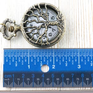 May include: An antique bronze pocket watch featuring a tree of life design. The watch face is visible. A blue ruler with centimetre and millimetre markings is placed below the watch. The watch is approximately 4 cm in diameter.