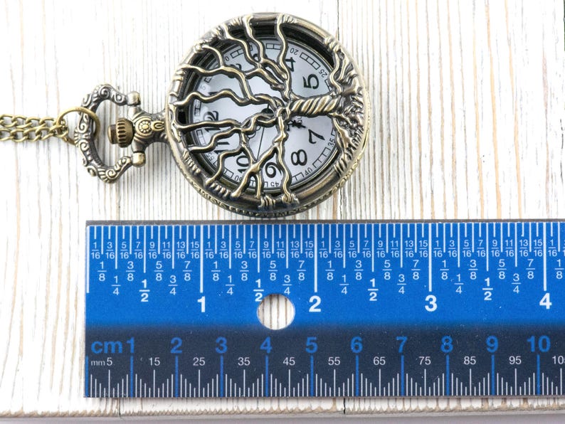 May include: An antique bronze pocket watch featuring a tree of life design. The watch face is visible. A blue ruler with centimetre and millimetre markings is placed below the watch. The watch is approximately 4 cm in diameter.