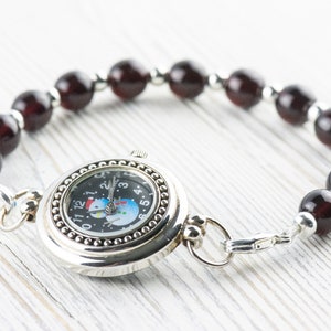 May include: A silver watch with a black face and a snowman design. The watch is attached to a bracelet made of dark red beads.