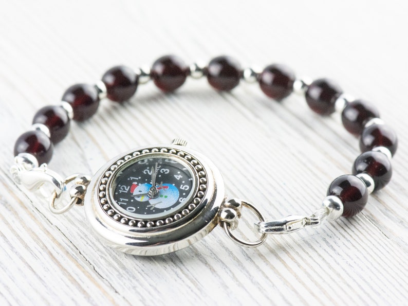 May include: A silver watch with a black face and a snowman design. The watch is attached to a bracelet made of dark red beads.